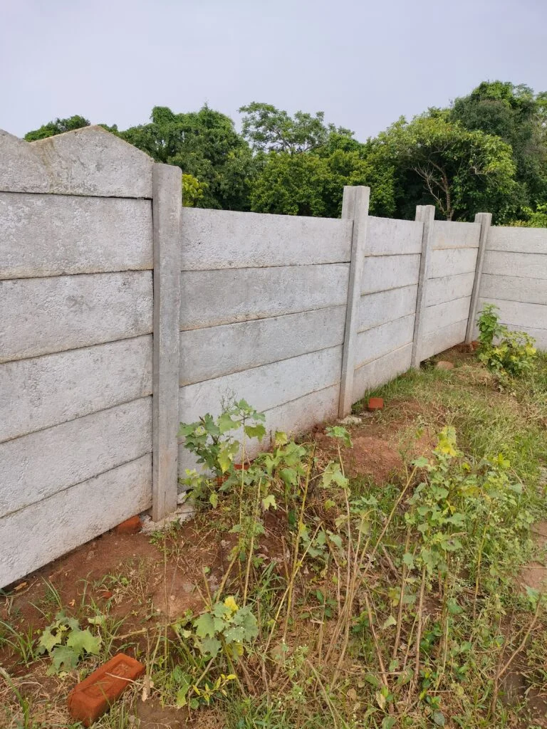 ckd enterprises A tall precast concrete boundary wall with smooth horizontal panels enclosing a garden area with wild green plants and some red bricks scattered on the ground.