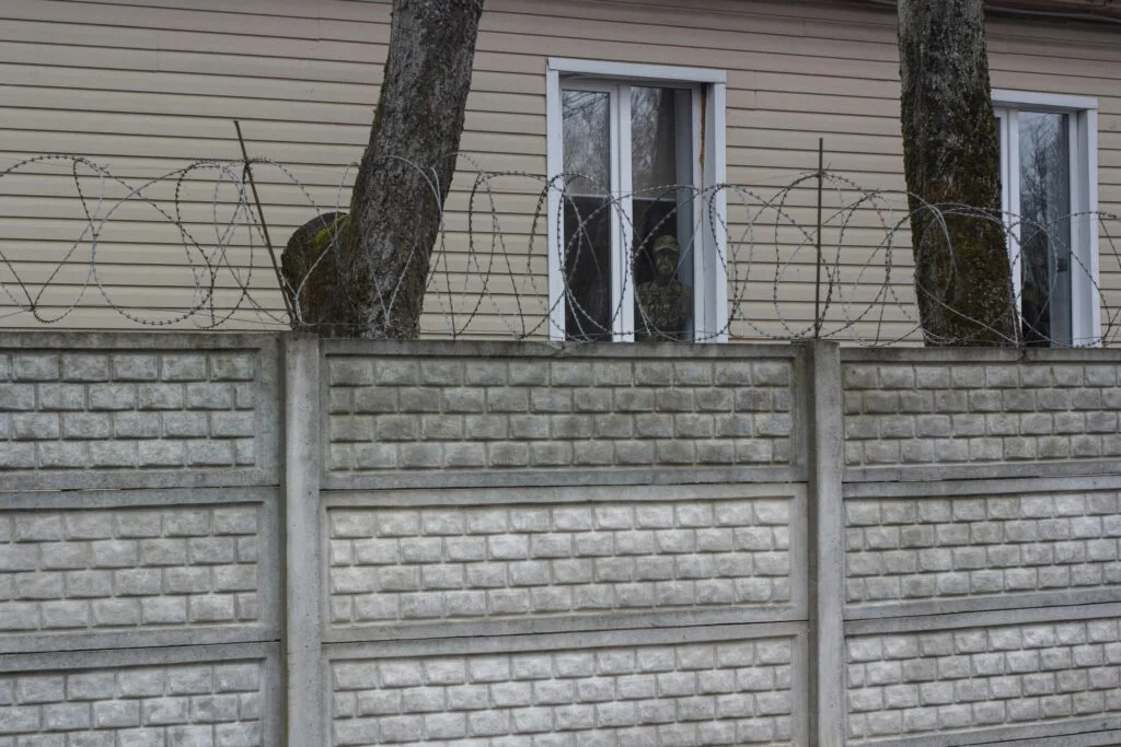 Concrete wall with barbed wire in front of a house with two windows.